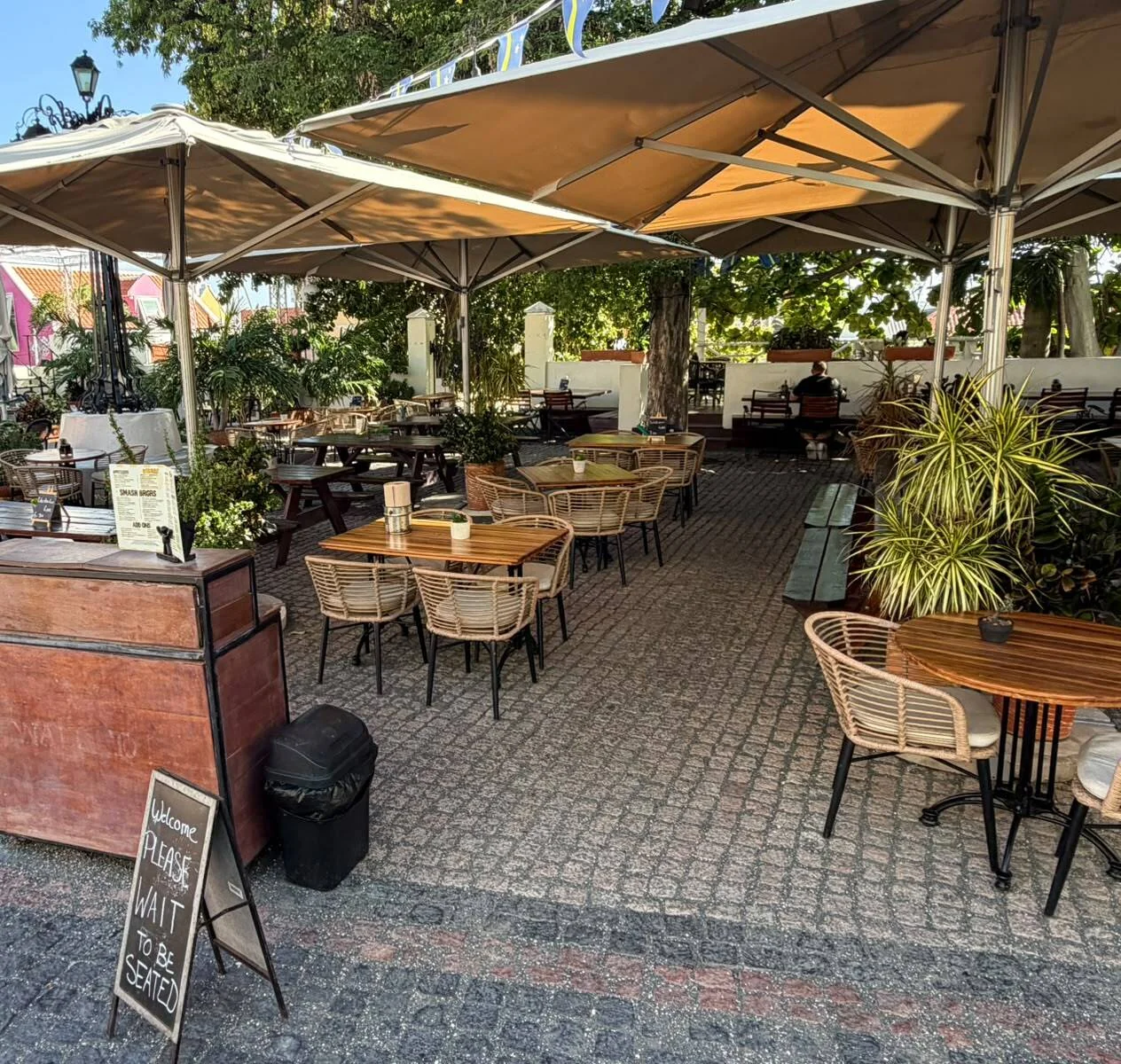 Freiluft-Speisebereich im Innenhof des BRGR Haus mit Rattanstühlen, Holztischen und großen Sonnenschirmen auf Kopfsteinpflaster im Kura Hulanda Village.