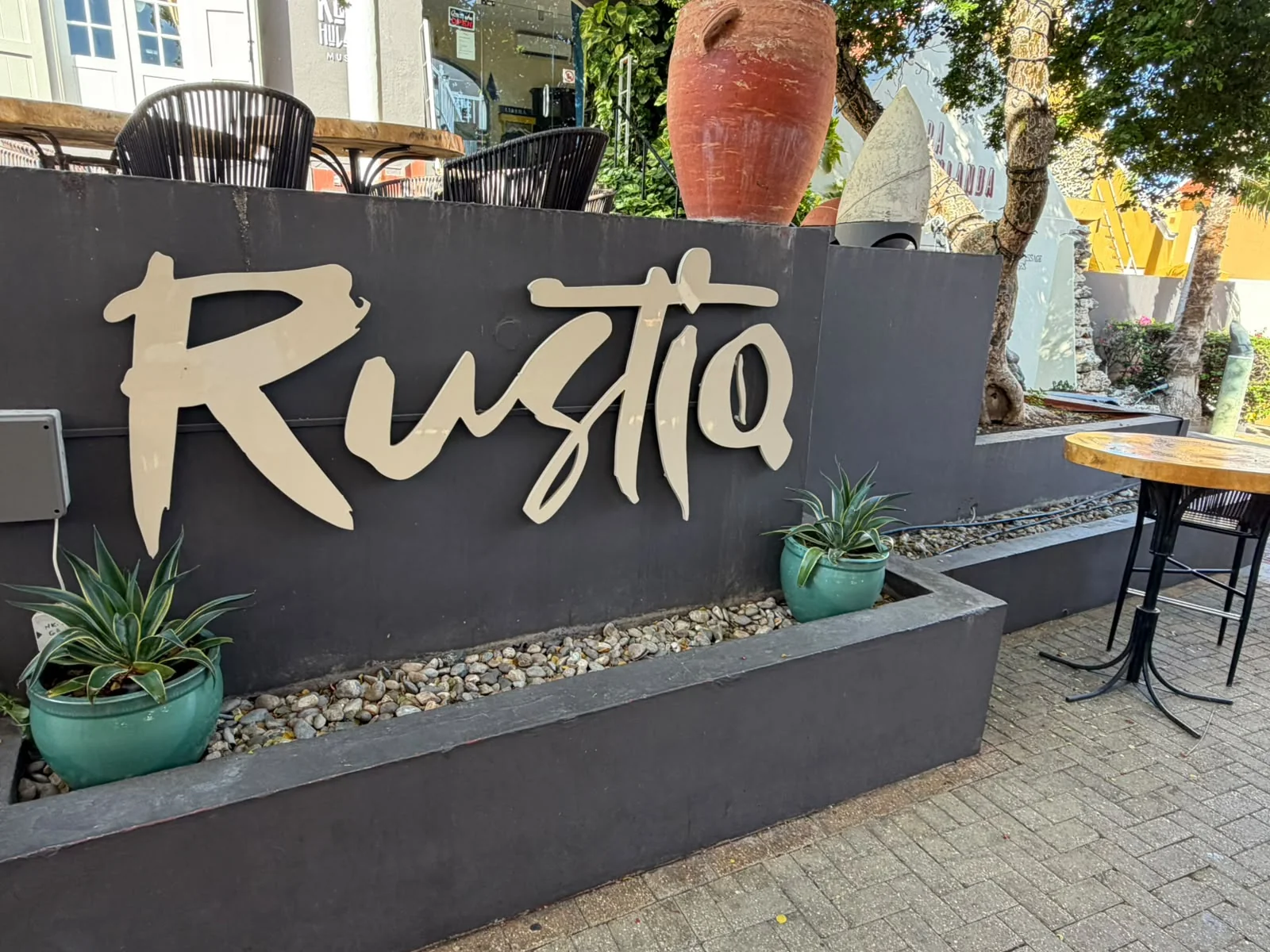 Das „Rustiq“-Namensschild mit cremefarbener Beschriftung an einer dunkelgrauen Pflanzwand am Eingang des Restaurants im Kura Hulanda Village.