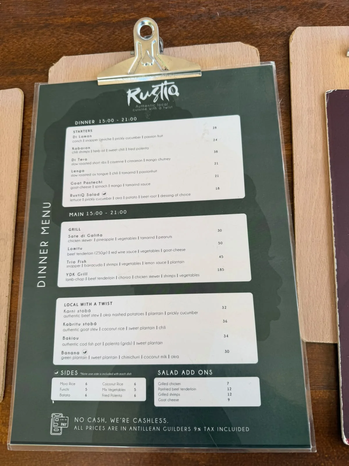 Speisekarte des Restaurants „Rustiq“ mit einer Auswahl an Vorspeisen, gegrillten Hauptgerichten und regionalen Spezialitäten, deren Preise in antillischen Gulden angegeben sind.