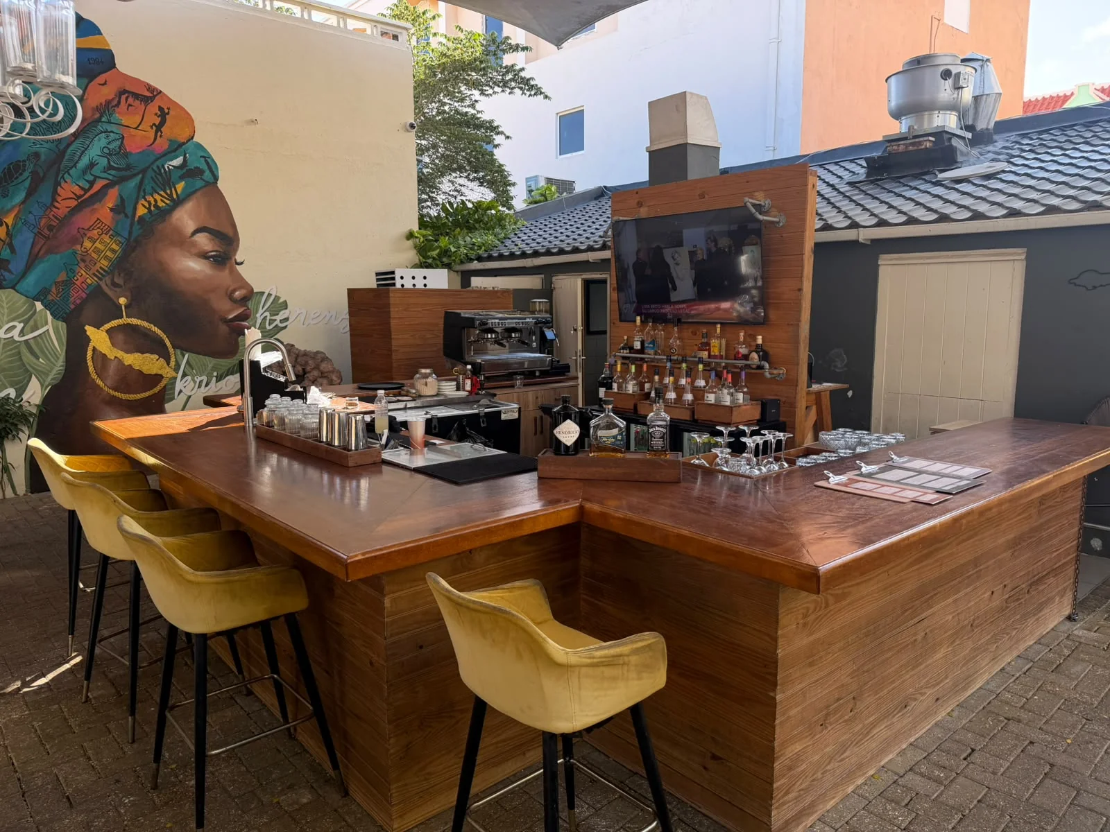 Die mit Spirituosen und Gläsern bestückte Open-Air-Bar von Rustiq mit einem an der Wand montierten Fernseher und dem Wandgemälde auf der Terrasse im Hintergrund.