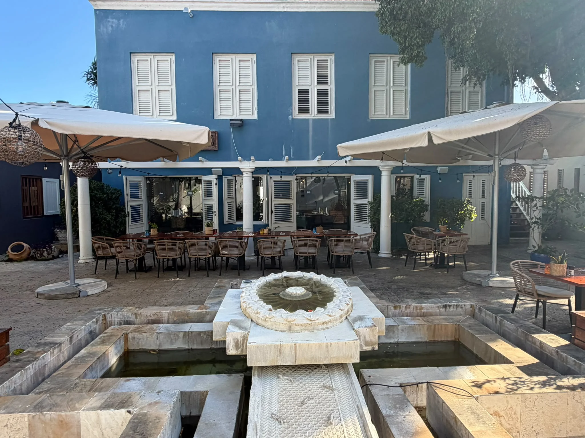 Die Terrasse im Innenhof der „Viva Pizza e Pasta Bar“ im Kura Hulanda Village mit einem mehrstufigen Steinbrunnen, cremefarbenen Sonnenschirmen und einem blauen Kolonialgebäude mit weißen