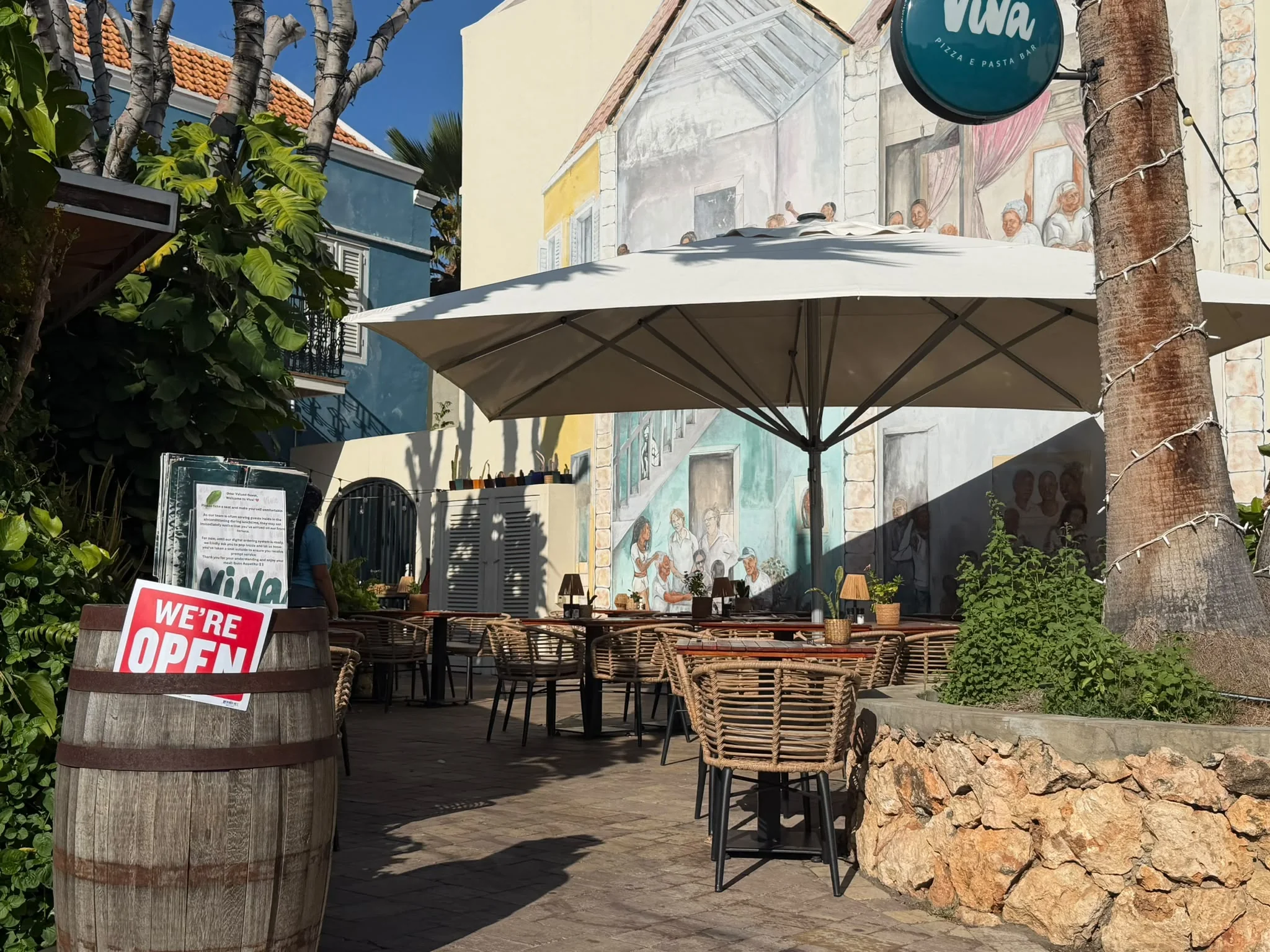 Eingang zur „Viva Pizza e Pasta Bar“ mit einem runden, blaugrünen Schild, einem Schild mit der Aufschrift „Wir haben geöffnet“, das auf einem Holzfass aufgestützt ist, und einer mit Wandmalereien