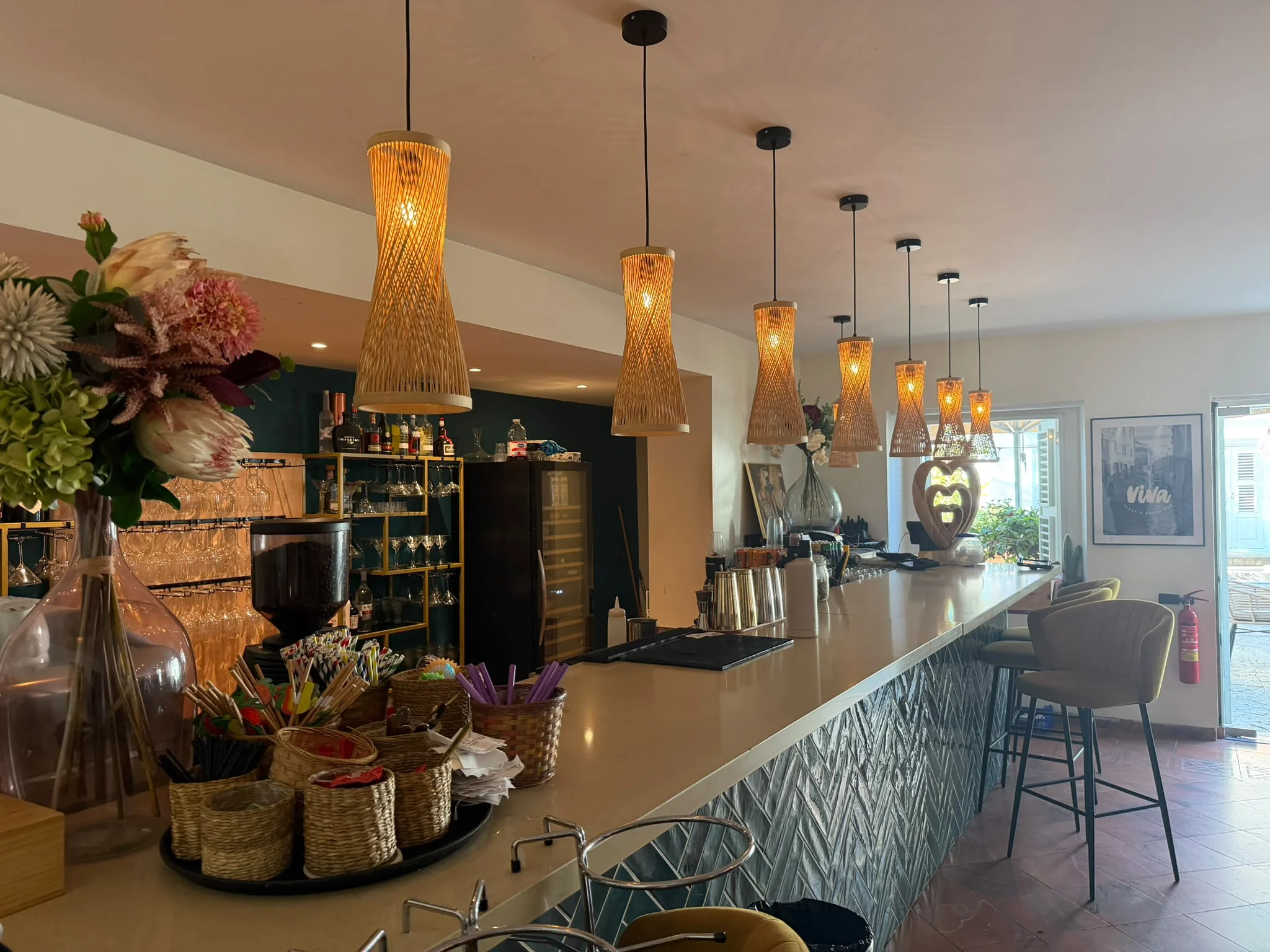 Theke in der „Viva Pizza e Pasta Bar“ mit geflochtenen Pendelleuchten, Barhockern und Regalen mit Glaswaren und Spirituosen.