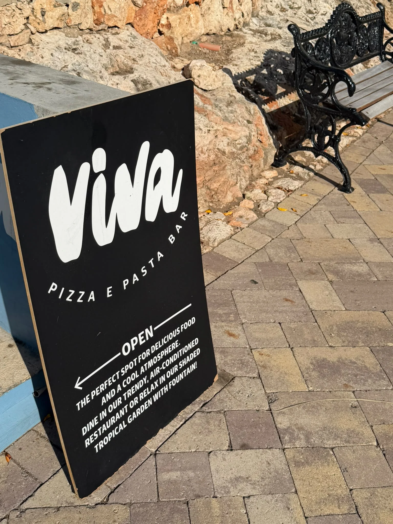 Schwarzes A-förmiges Gehwegschild für die „Viva Pizza e Pasta Bar“, das für den klimatisierten Speisesaal und den schattigen Garten mit Springbrunnen wirbt.