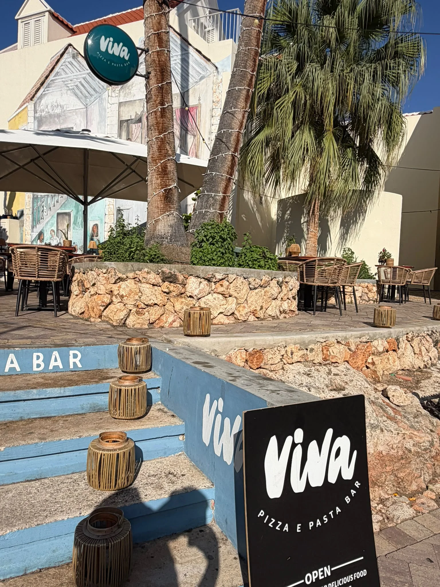 Blau gestrichene Stufen und Palmen am Eingang zur Straße der „Viva Pizza e Pasta Bar“, über dem das runde Schild des Restaurants hängt.